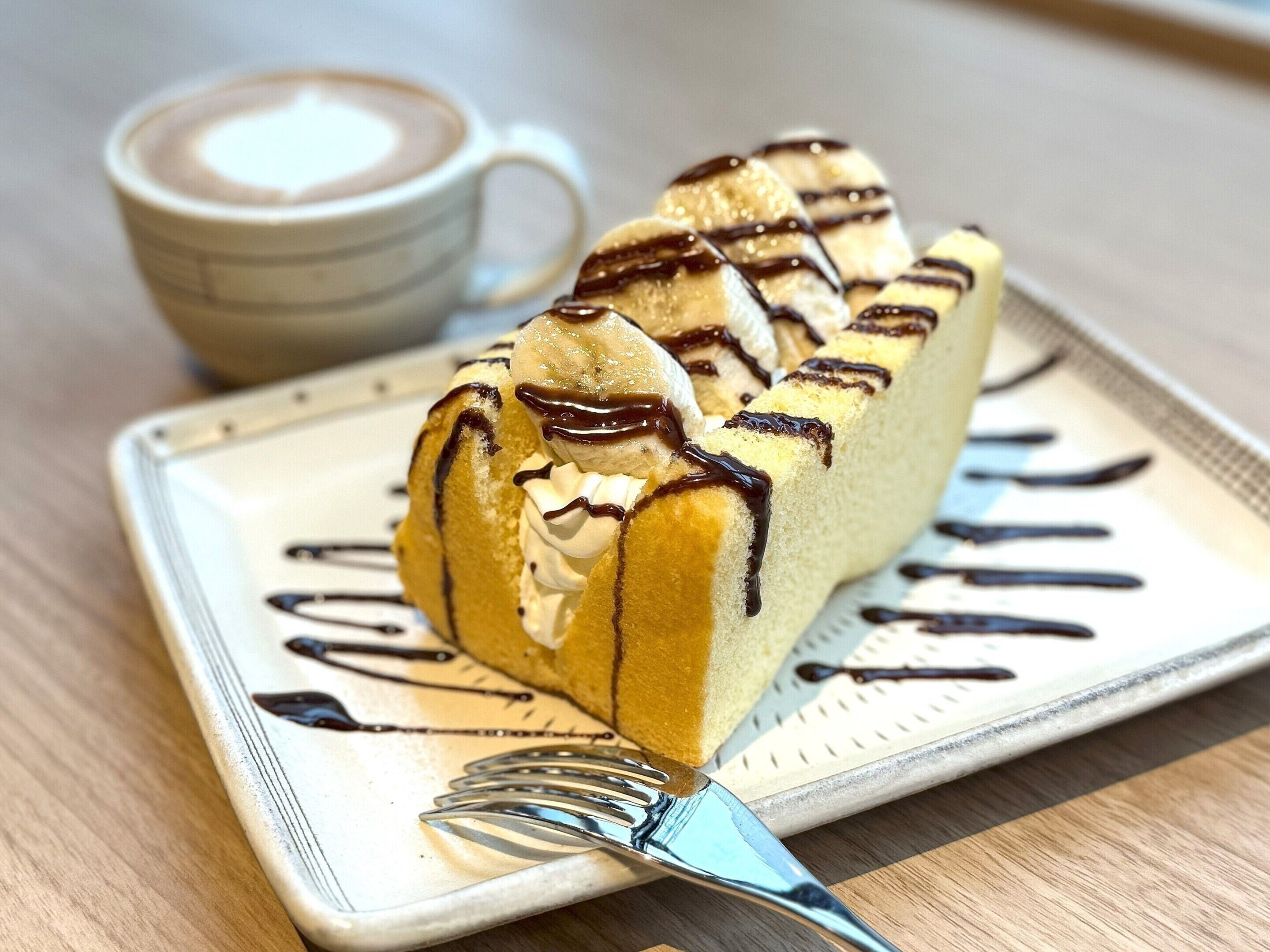 Una porción de pastel con crema batida y un chorrito de chocolate sobre un plato, acompañada de una taza de café.
