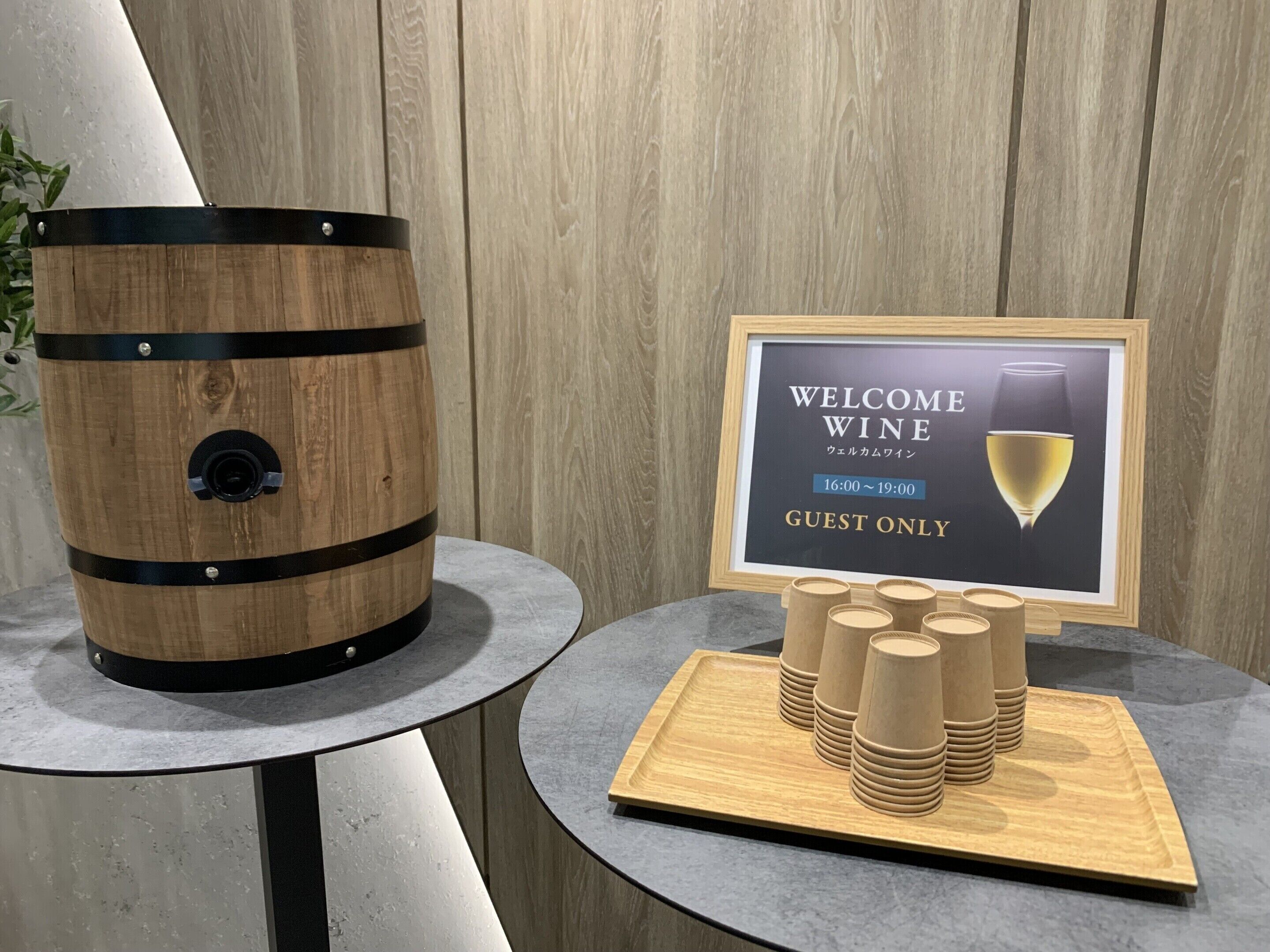 Un barril de madera, un cartel enmarcado con el texto 'WELCOME WINE' y vasos de papel apilados sobre una mesa.