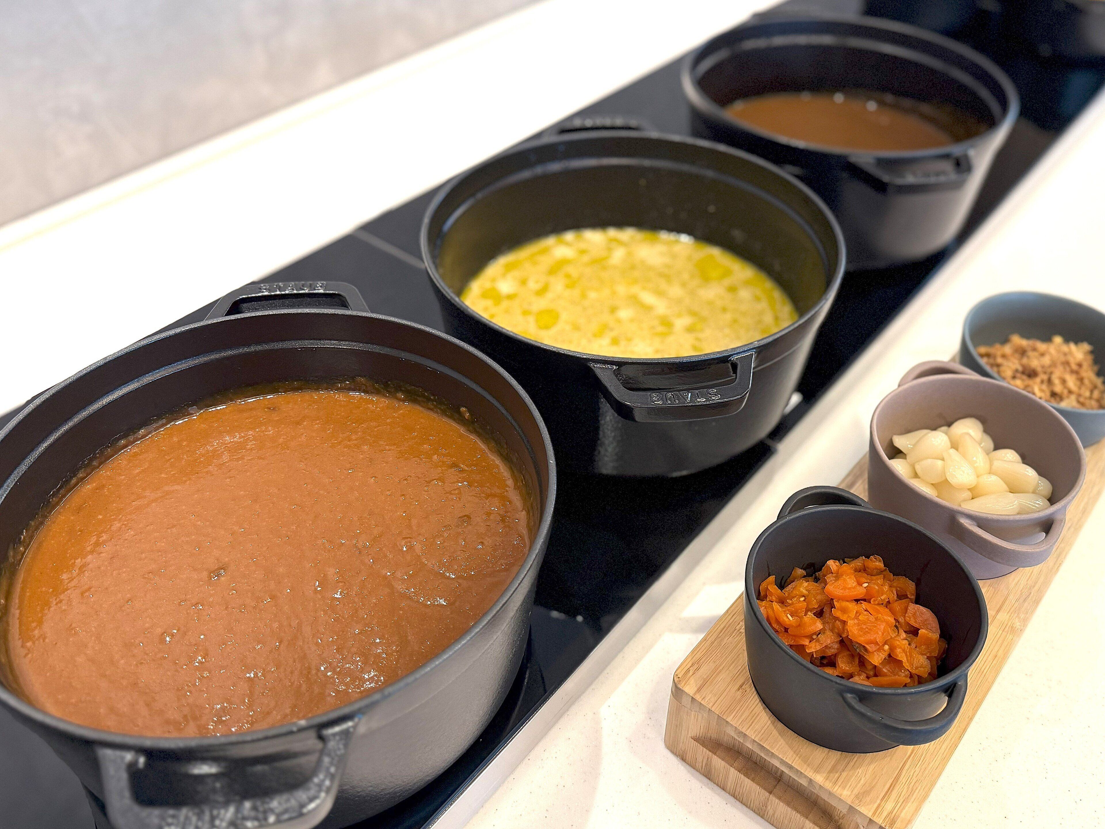 Quatre casseroles sur une cuisinière, chacune contenant des aliments différents, et trois petits bols remplis de divers ingrédients, le tout disposé sur une planche en bois.