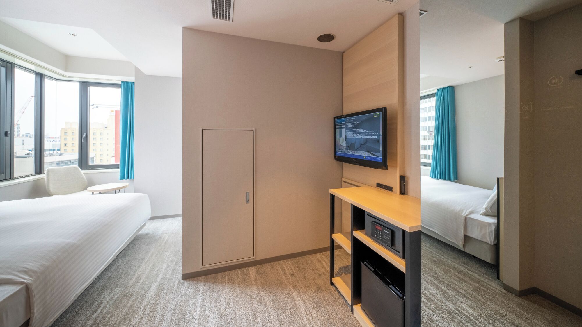 Quarto de hotel com cama, escrivaninha com televisão, guarda-roupa e vista da cidade através de amplas janelas.
