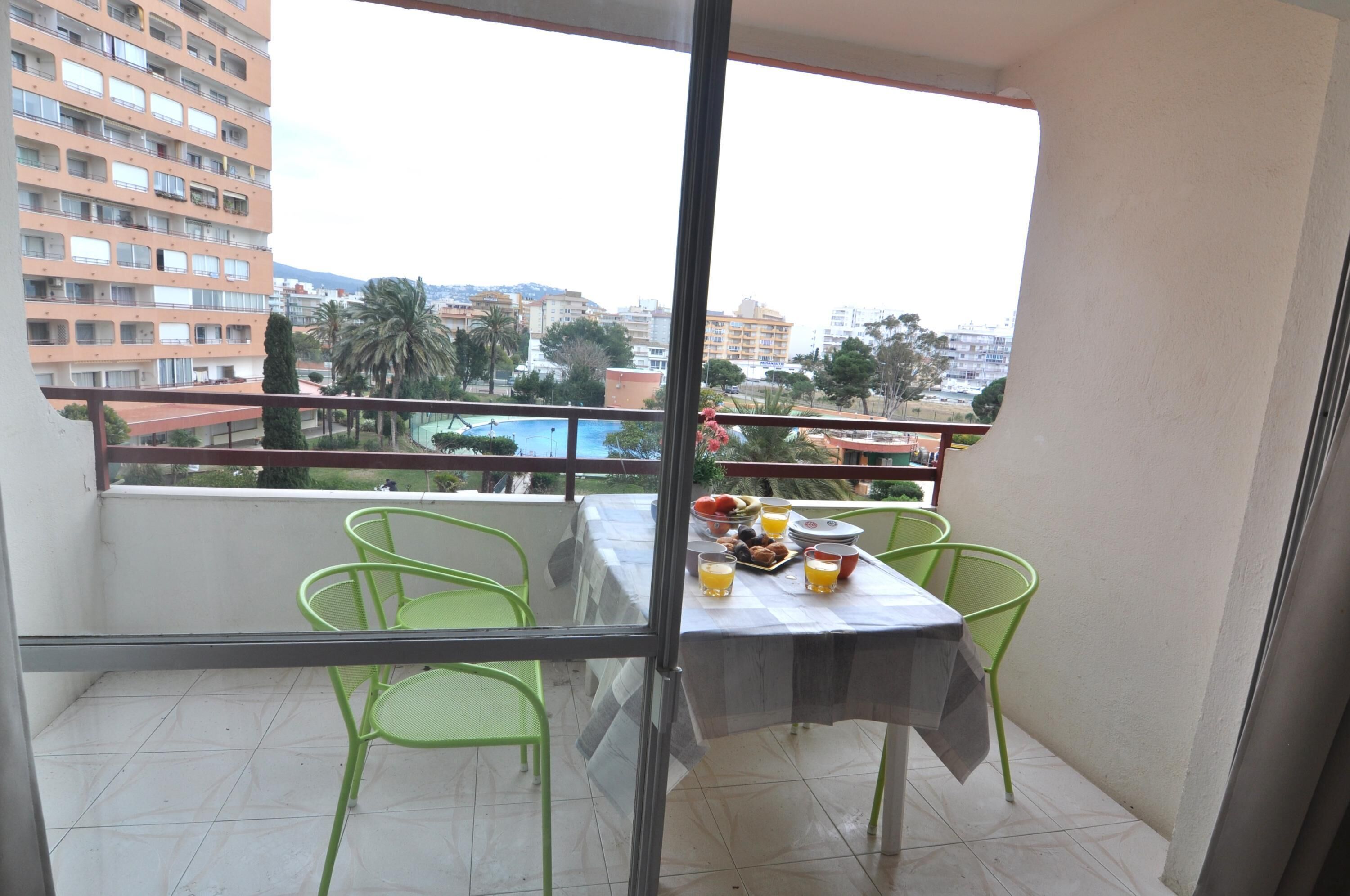 A balcony with a table set for breakfast, green chairs, and a view of a cityscape with buildings and palm trees.