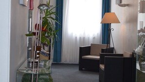 A modern hotel room with a glass partition, a potted plant, a sofa, a lamp, and a view of the outdoors through a window with blue curtains. - Hotel de La Croix de Malte (Cherbourg-en-Cotentin)