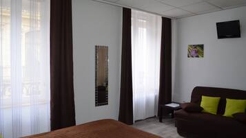 Een hotelkamer met een bed, een bruine bank met gele kussens, een kleine tafel, een aan de muur bevestigde televisie en een raam met dunne gordijnen.