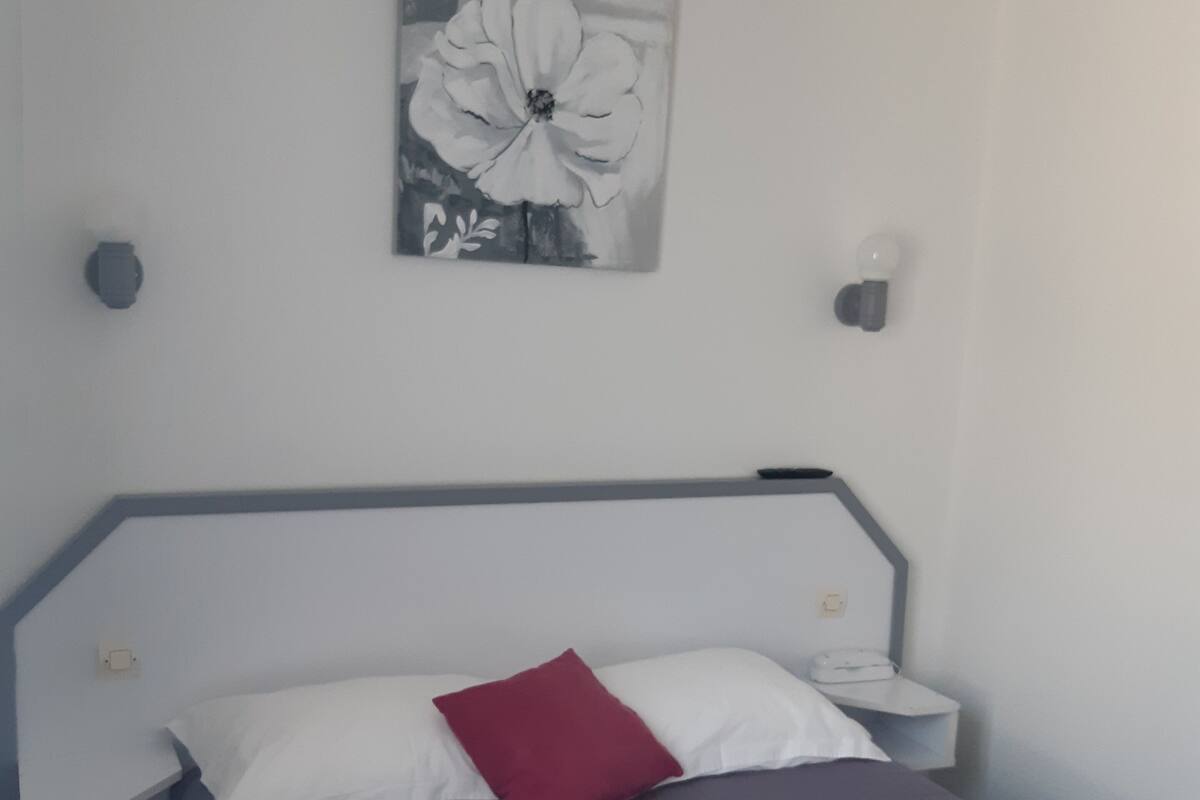 A bedroom with a bed, a nightstand, a red pillow, a wall-mounted lamp, and a painting of a flower.
