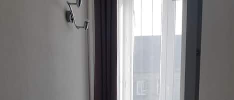 Una habitación pequeña y tenue, con una sola silla gris, una mesita con un objeto rojo y una ventana con cortinas.