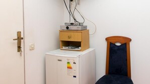 Una habitación compacta con un refrigerador blanco, un pequeño mueble de madera, un televisor montado en la pared y una silla azul oscuro.