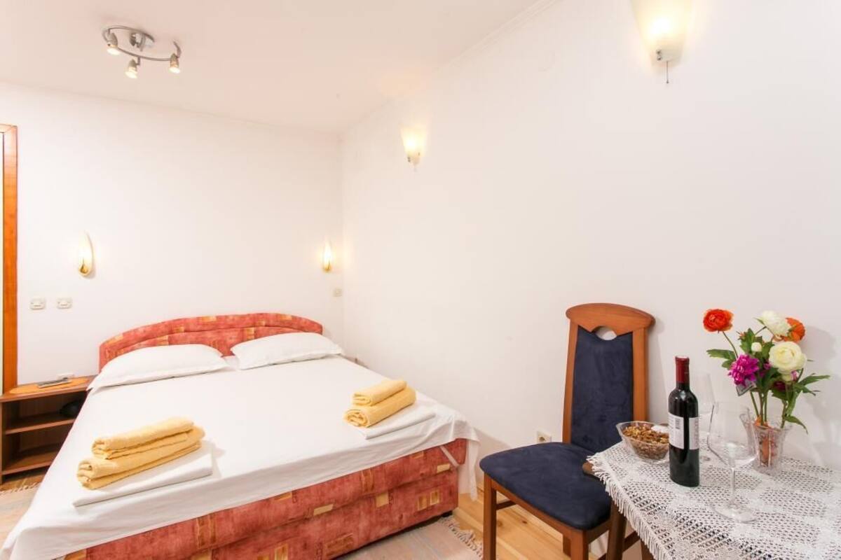 Ein Schlafzimmer mit Bett, Nachttisch, Stuhl und einem kleinen Tisch mit Flasche und Blumen.