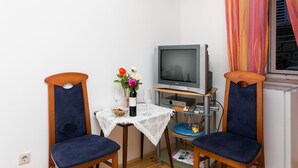 Una habitación pequeña con televisión, dos sillas, una mesa con un jarrón de flores y una ventana con cortinas.