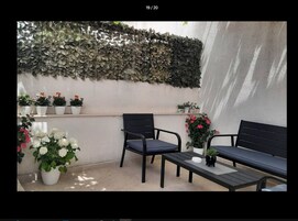 Um espaço ao ar livre com paredes cobertas de folhagem, vários vasos de plantas e duas cadeiras pretas com uma pequena mesa.