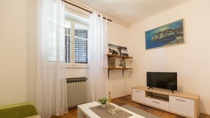 Un salón con una mesa blanca, un televisor sobre un soporte de madera, una ventana con cortinas blancas y un cuadro en la pared.
