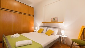 Una cama bien hecha con almohadas blancas y amarillas, una colcha verde y una toalla blanca a un lado. Se observa un armario de madera, una mesita de noche con una lámpara y una silla con cojín amarillo.