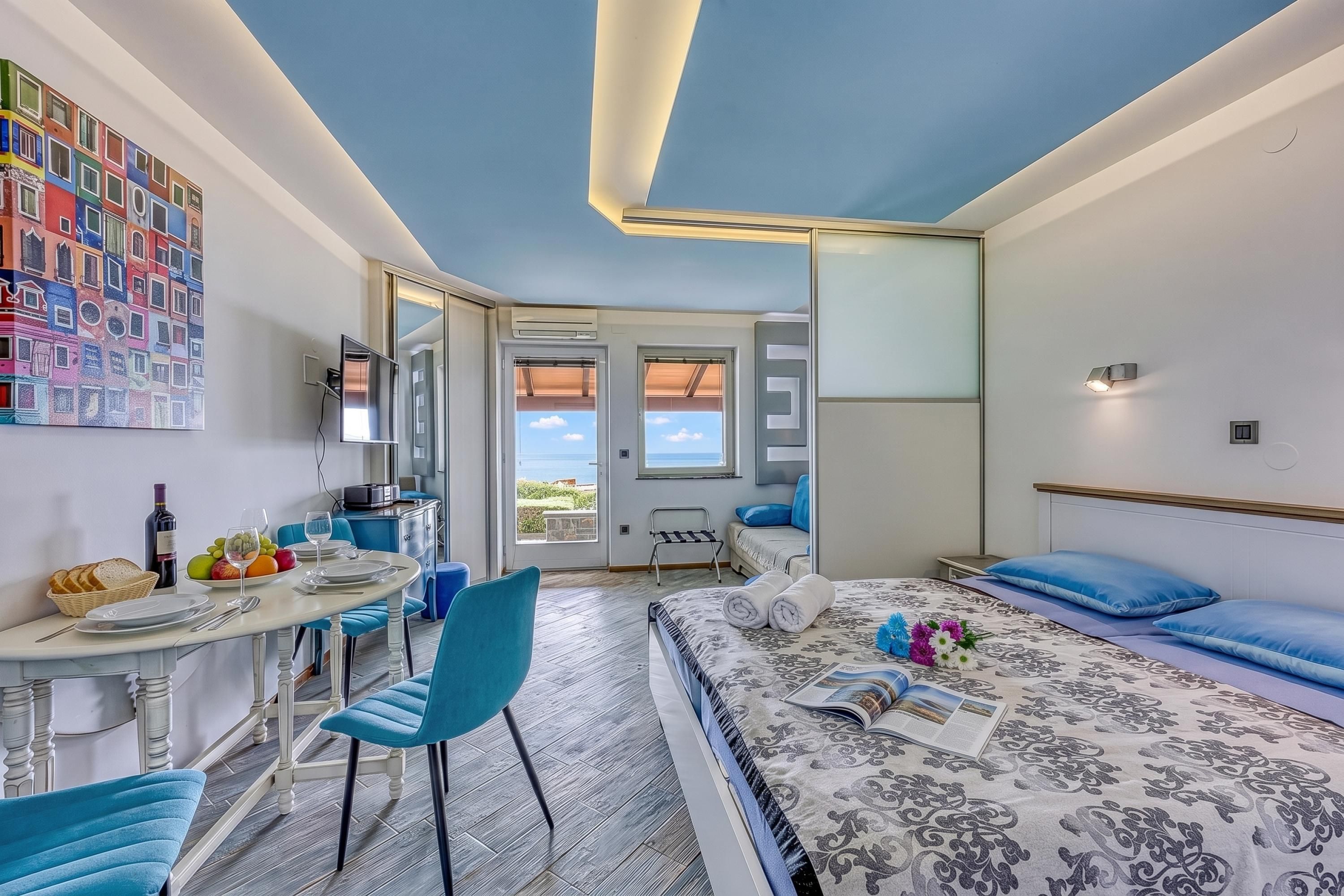 Habitación de hotel con cama, una mesa de comedor con sillas, una pequeña cocina y un balcón con vistas.