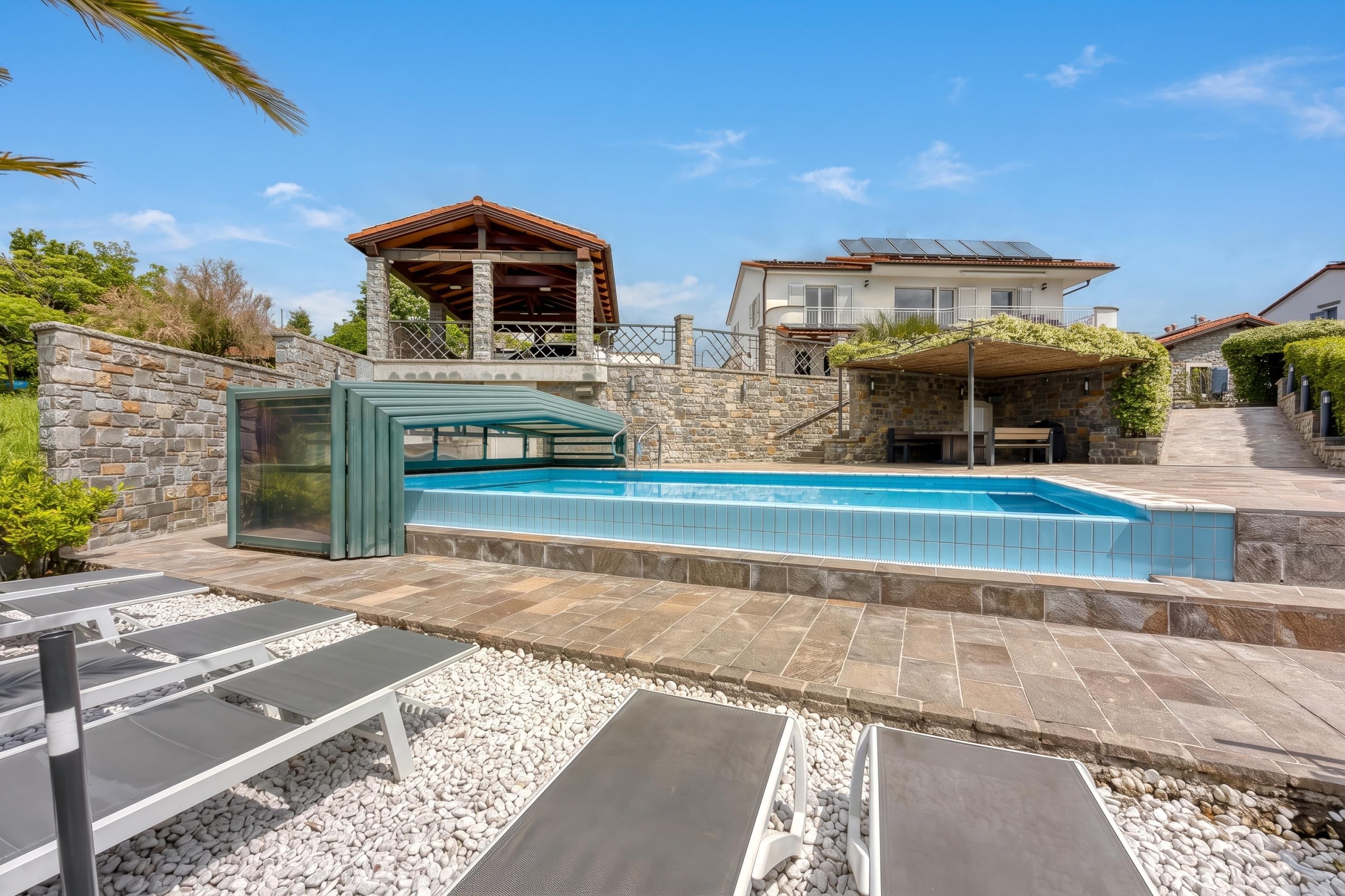 A swimming pool with lounge chairs, a stone wall, and a house with a balcony.