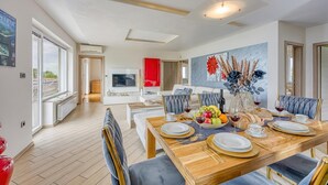 A modern dining area with a wooden table set for six, a television in the background, and a large window with a view. - Pool Apartments Debeli Rtič (Ankaran)