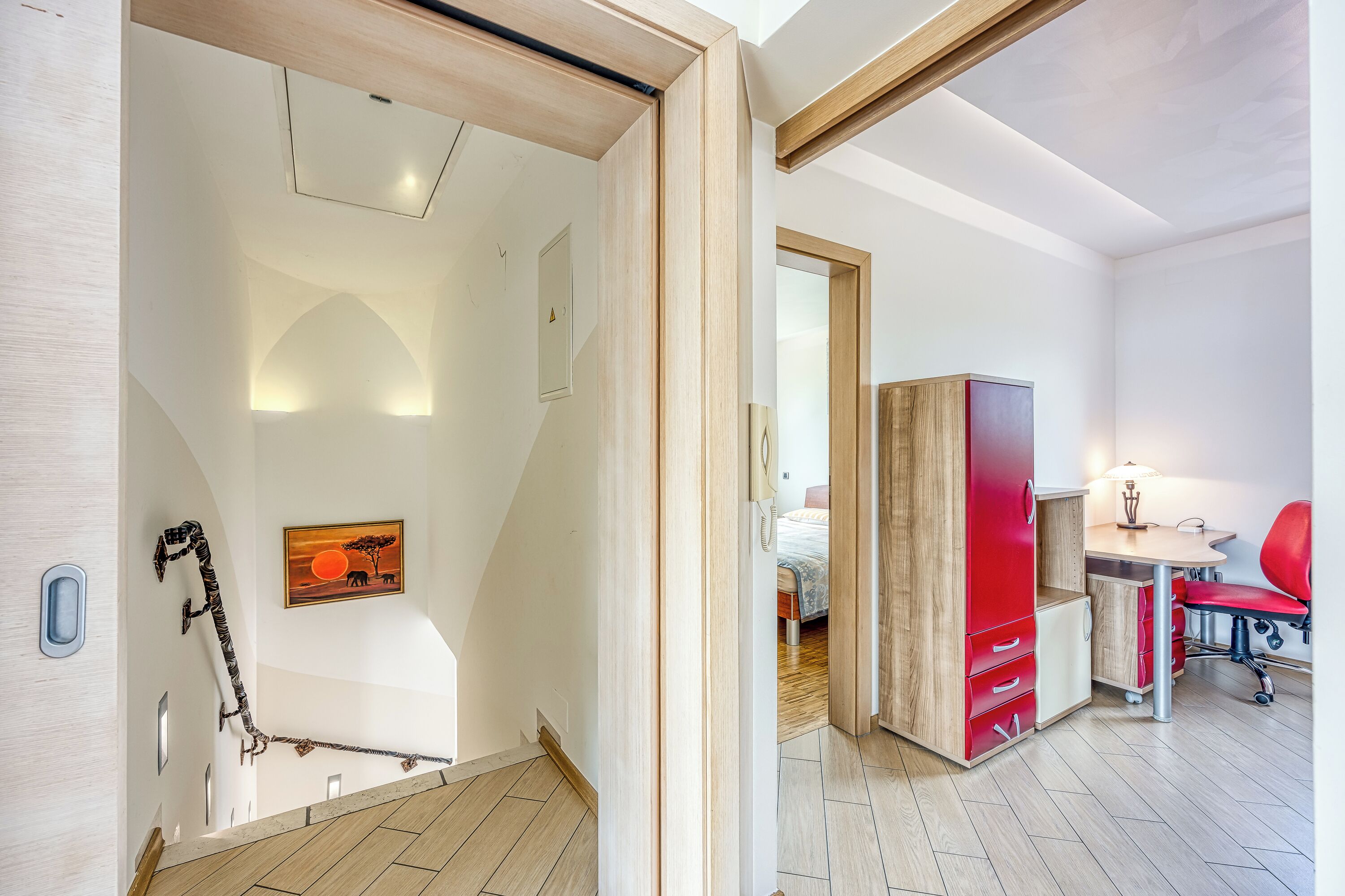 A modern bedroom with a wooden staircase, a desk with a chair, and a bed with a red headboard.