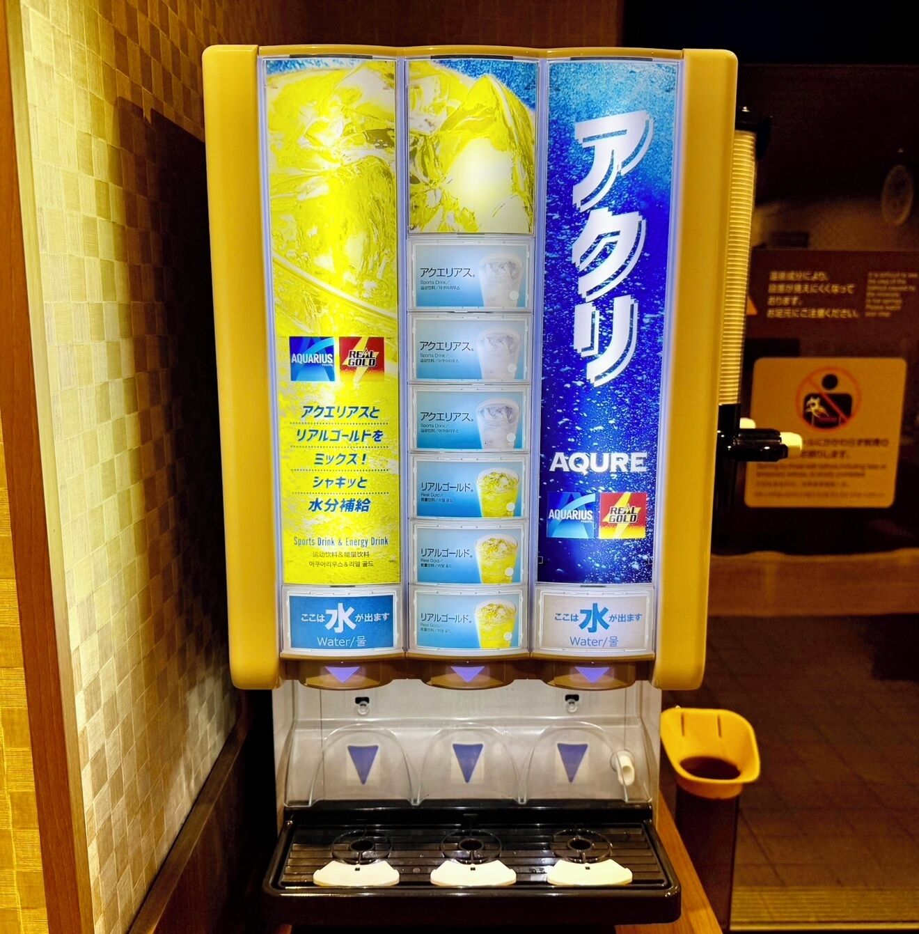 A water dispenser with multiple drink options, including sports drinks and energy drinks, and a sign indicating the brand 'Aquarius' and 'Real Gold'.