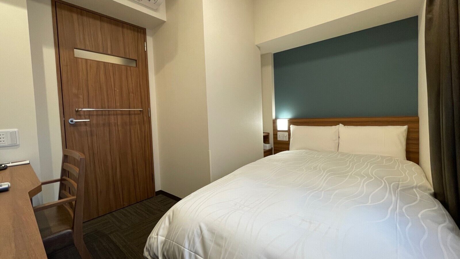 A hotel room with a bed, a wooden door, a desk, and a chair.