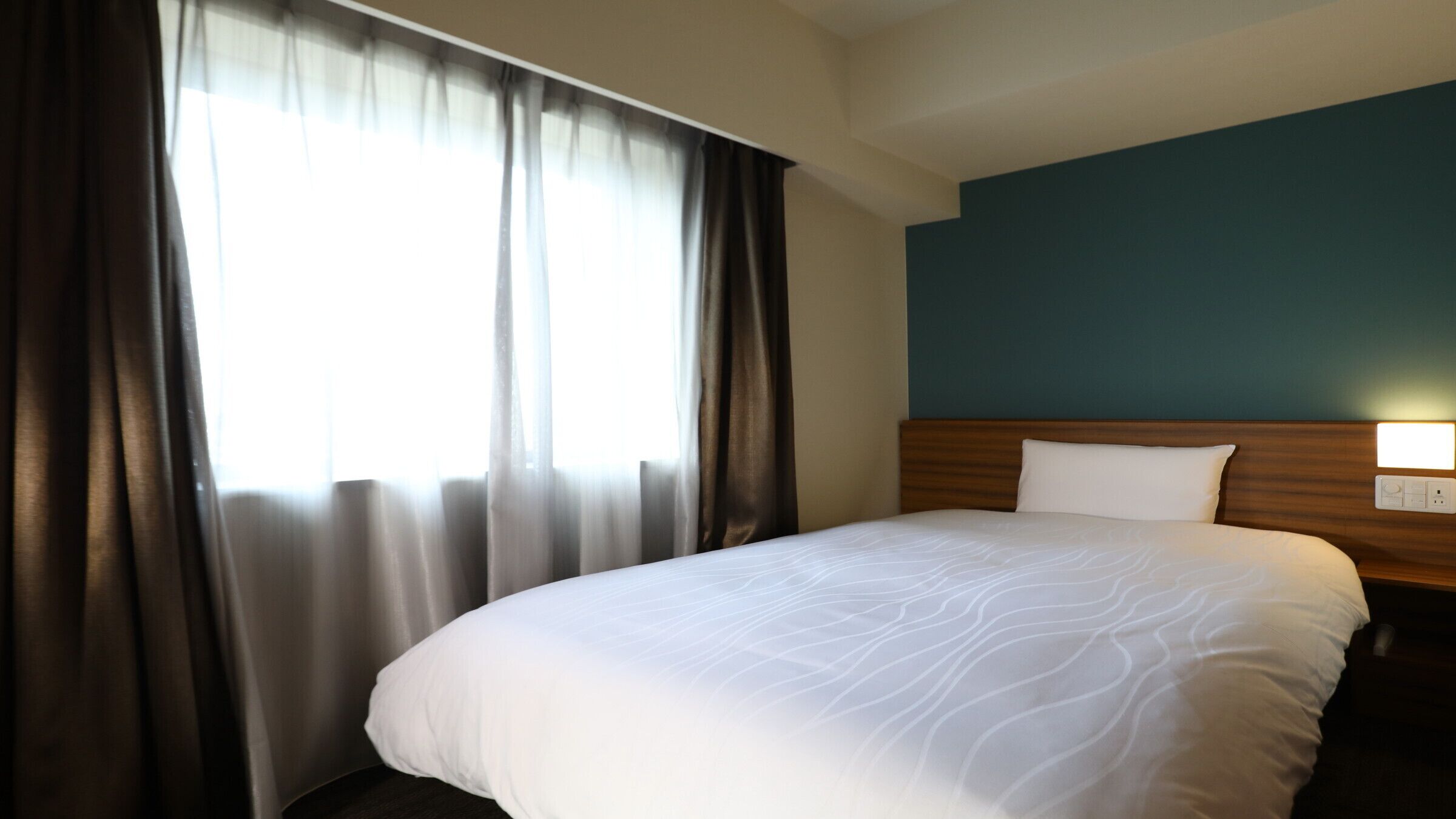 A hotel room with a large bed, a wooden headboard, a window with curtains, and a wall-mounted light.