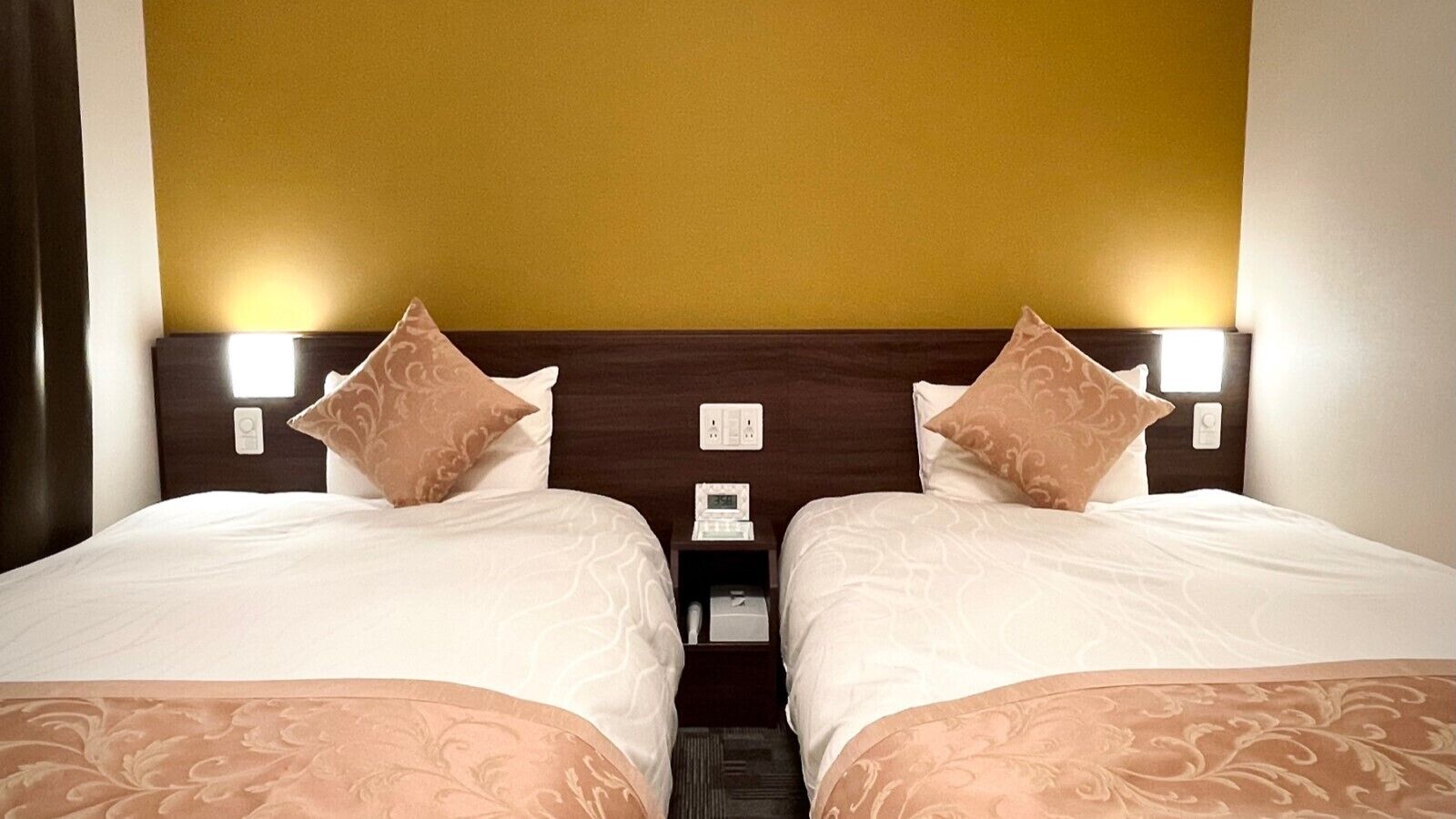 A hotel room with two beds, a wooden headboard, bedside lamps, a nightstand, and a wall socket.