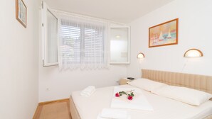 A neatly made bed with white linens, a bedside table with a lamp, and a window with blinds. - Apartment Marina (Dubrovnik)