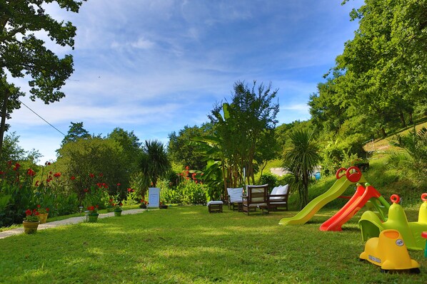 A backyard with a grassy lawn, a wooden bench, a small table, and a children's play area with slides.