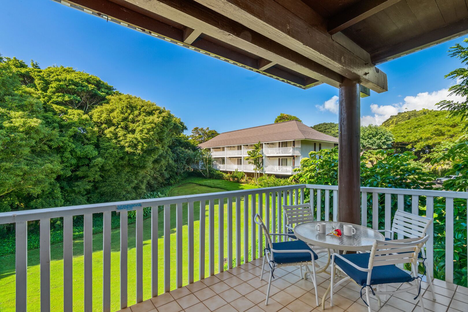 A balcony with a table and chairs, overlooking a lush green landscape and a two-story building.