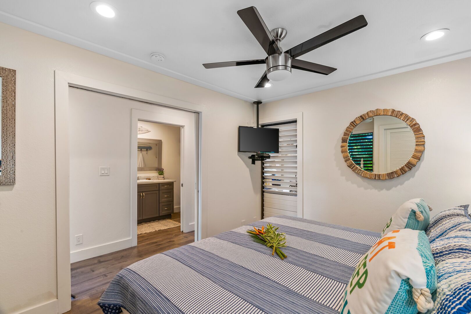 A bedroom with a bed, a ceiling fan, a TV, and a bathroom visible through an open door.