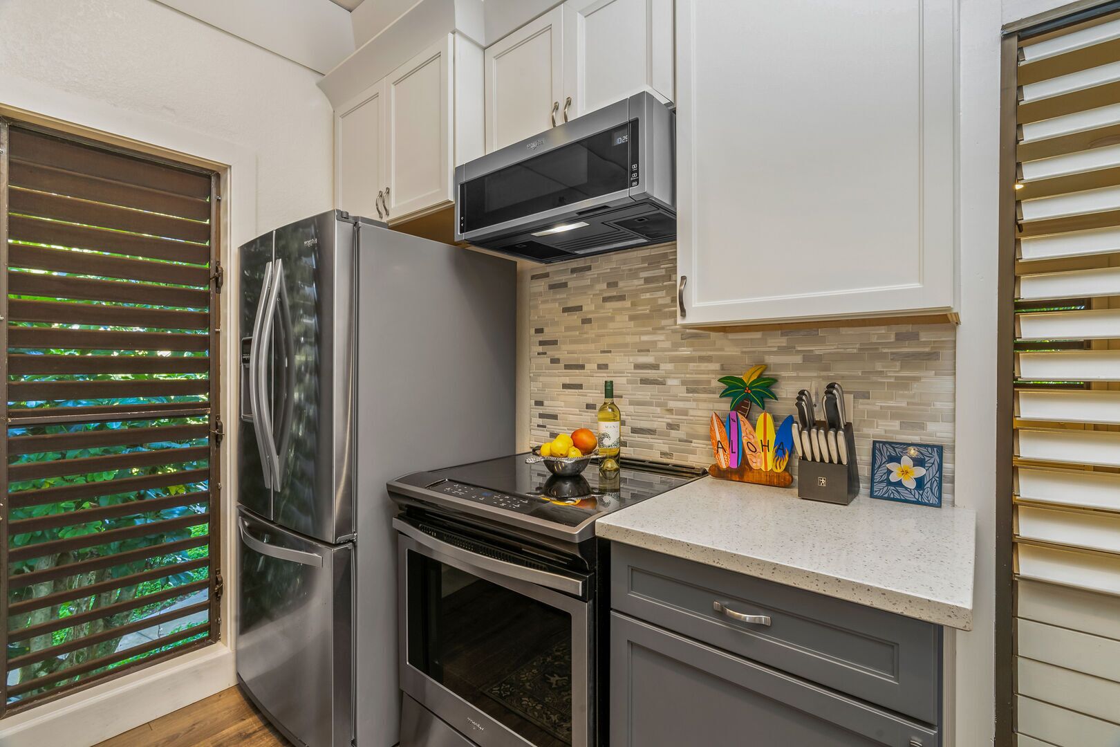 A compact kitchen with a refrigerator, oven, microwave, and a small countertop with a fruit bowl, wine bottle, and kitchen utensils.