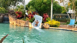 A swimming pool with a water slide, surrounded by tropical plants and rocks.