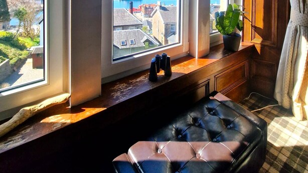 Una habitación con tres grandes ventanales que dan a una vista costera, un banco negro tapizado y una planta en maceta en el alféizar.