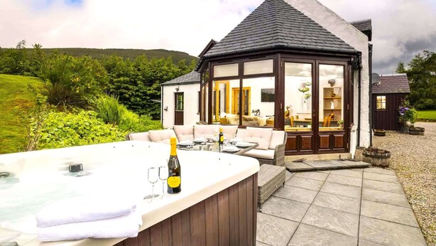 A cozy outdoor seating area with a hot tub, a bottle of champagne, and a table set for two. - Rowan Cottage - Retreat Group (Aviemore)