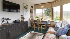 A modern living room with a sofa, a coffee table, a TV, and a dining area with chairs and a table. - Rowan Cottage - Retreat Group (Aviemore)