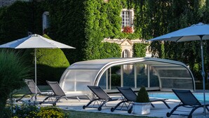 A greenhouse structure with a glass roof, surrounded by outdoor seating with umbrellas, a pool, and well-maintained greenery. - Les Granges du Château : Charming gîte with swimming pool in Burgundy (Chevannes)