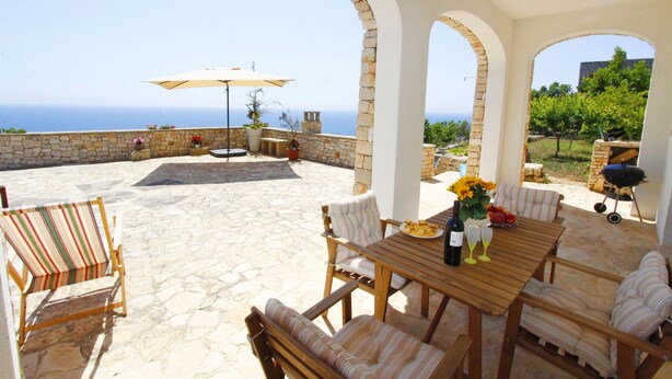 Una terrazza esterna con pavimento in pietra, arredi in legno e vista sul mare.