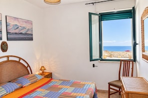 A bedroom with a bed, a chair, a table, and a window with a view of the sea.