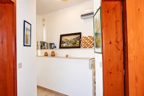A hallway with a wooden door, a white wall, a shelf with books and pottery, and a framed painting.