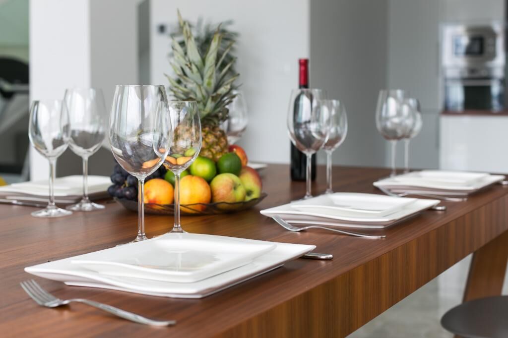 Une table à manger dressée avec de l’assiette blanche, des couverts et des verres à vin vides, avec un panier de fruits et une bouteille en arrière-plan.