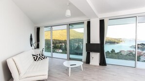 Ein modernes Wohnzimmer mit einem weißen Sofa, einem kleinen weißen Tisch und einem großen Fenster mit Blick auf eine Küstenlandschaft.