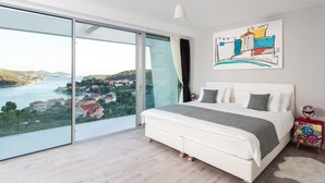 Ein Schlafzimmer mit einem großen Bett, Blick auf eine Küstenlandschaft und einem farbenfrohen Kunstwerk an der Wand.