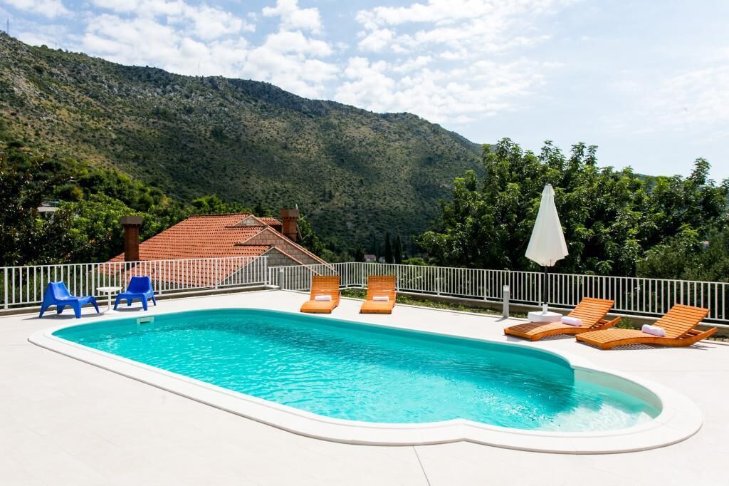 Piscina com espreguiçadeiras e um guarda-sol branco, cercada por uma grade branca e um cenário montanhoso.