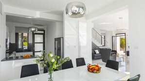 A modern kitchen with a central island, a dining area with a white table and chairs, and a staircase leading to an upper level. - Villa Andromeda Sea Caves (Paphos)