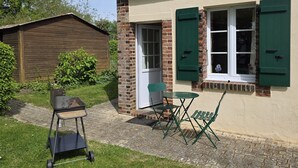 Una casa con fachada de ladrillo, contraventanas verdes, un pequeño patio con mesa y sillas, y un jardín.