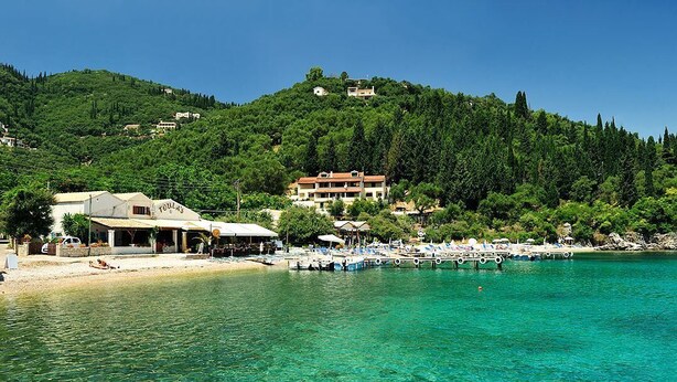 A coastal area with a clear blue sea, a sandy beach, and a hill with buildings. - The Fisherman's Cottage (Corfu)