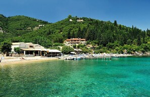 A coastal area with a clear blue sea, a sandy beach, and a hill with buildings. - The Fisherman's Cottage (Corfu)