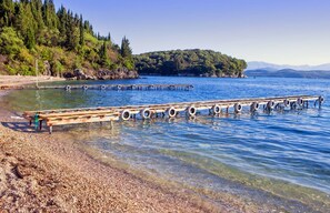 A wooden pier extending into a clear lake, surrounded by pebbles and trees. - The Fisherman's Cottage (Corfu)