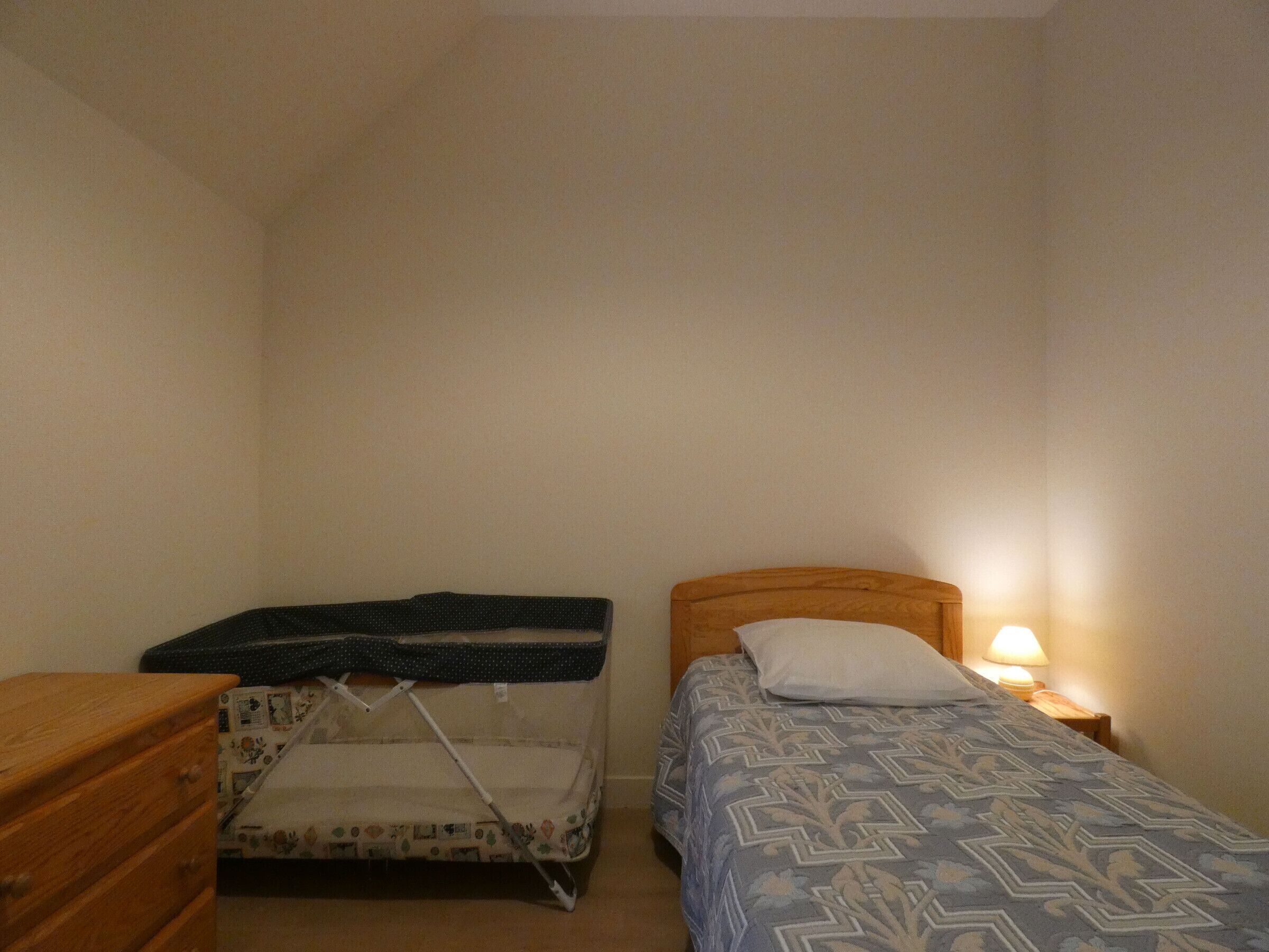 A small, dimly lit room with two single beds, one with a polka-dotted cover and the other with a patterned bedspread. A wooden dresser is on the left, and a small table with a lamp is on the right.