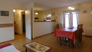 Un comedor con mantel rojo, sillas de madera, piso de baldosas y una cocina al fondo.