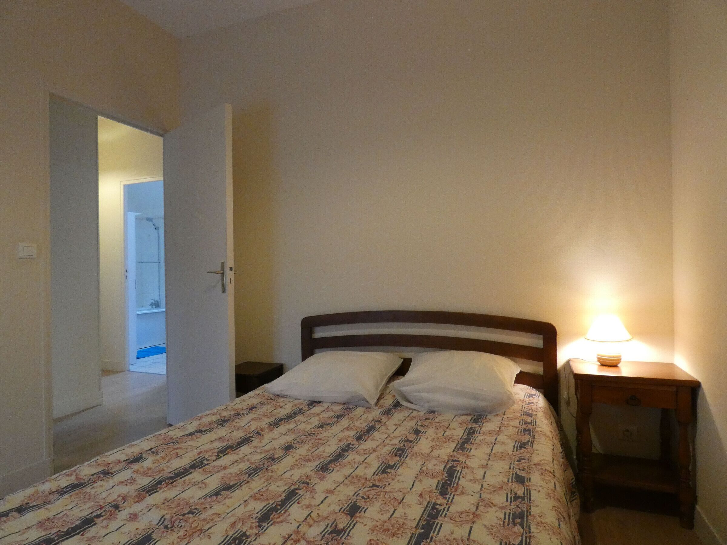 Un dormitorio con cama, mesita de noche y lámpara. Hay una puerta que da a un baño.