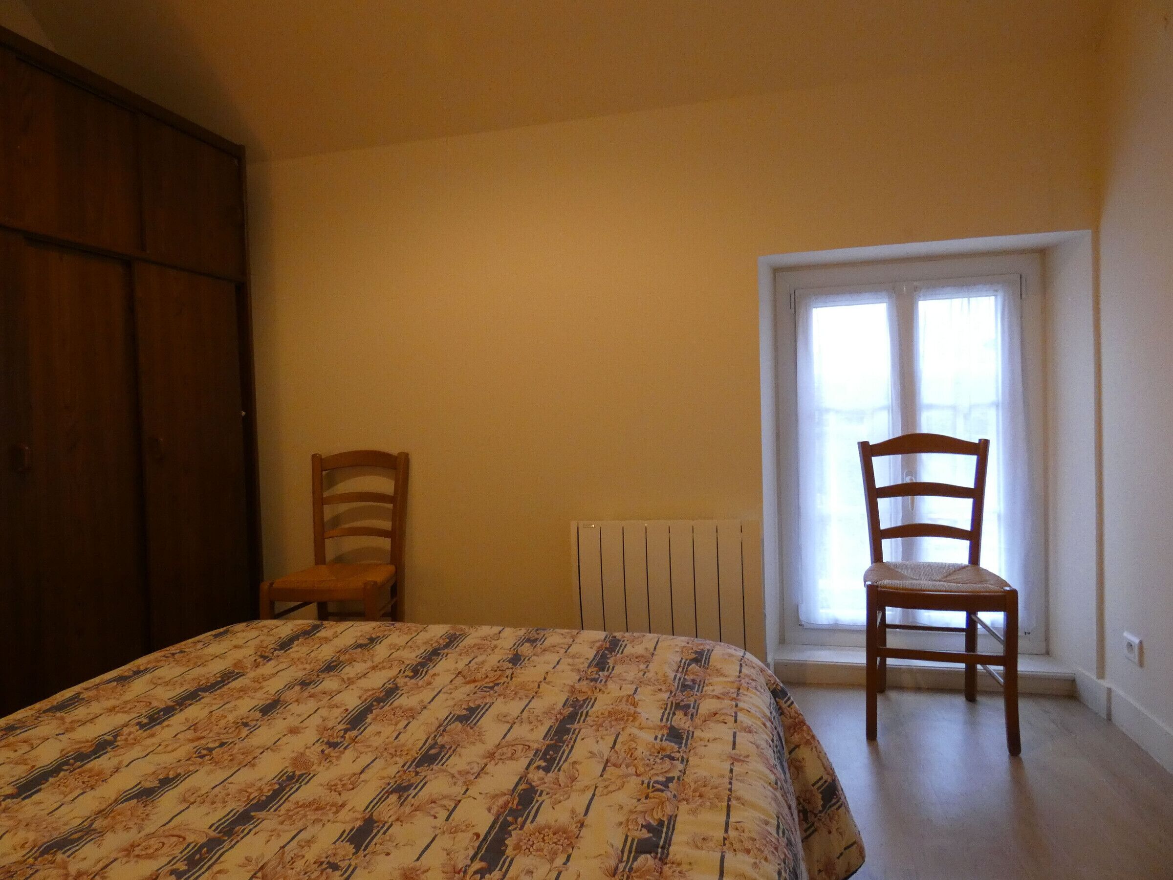 Une chambre à coucher avec un lit, deux chaises en bois, un radiateur et une fenêtre avec des rideaux.