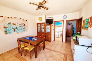 Un comedor con una mesa y sillas de madera, un ventilador de techo, un televisor montado en la pared y vistas a la playa a través de una puerta abierta.